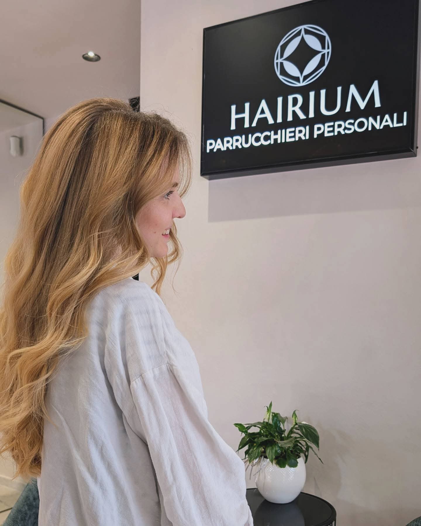 Cliente con lunghi capelli biondi mossi accanto all'insegna Hairium